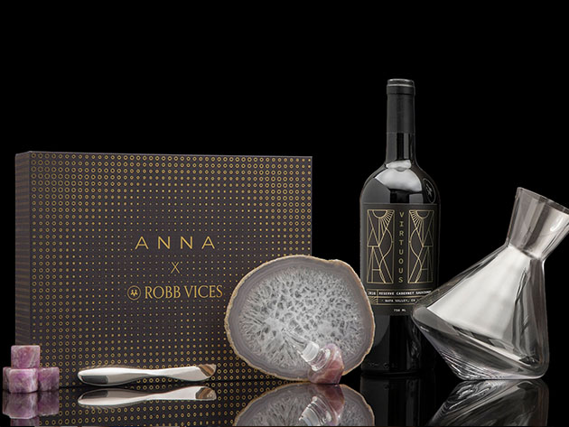 Una caja que dice "ANNA X ROBB VICES," junto a una botella de vino, piedra decorativa y jarra de cristal 