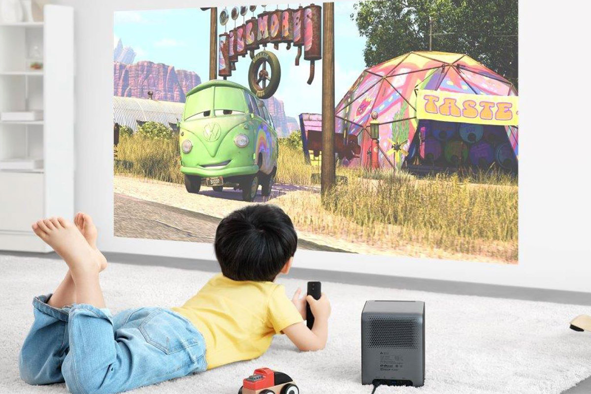 a little boy in a yellow shirt and jeans lays on a carpet watching a Cars movie that is projected on the wall